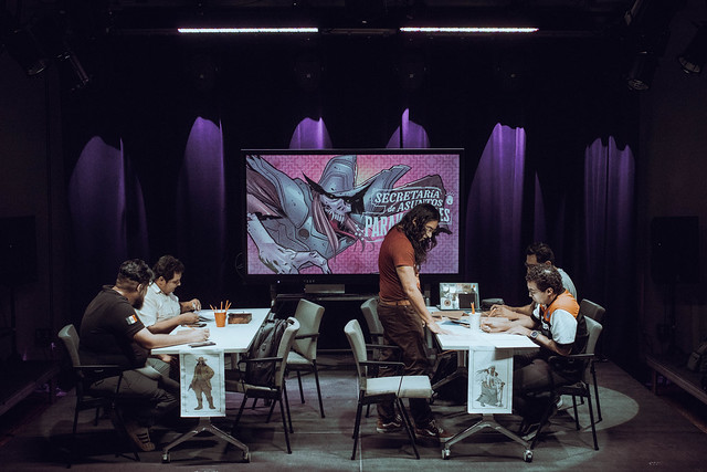 Dos mesas con varias personas sentadas alrededor mientras juegan una partida de rol. Al fondo hay una pantalla con una ilustración del juego 'Secretaría de Asuntos Paranormales' desarrollado por la comunidad Leyendas del Noreste del Laboratorio Cultural Ciudadano de Monterrey (México).