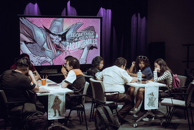 Dos mesas con varias personas sentadas alrededor mientras juegan una partida de rol. Al fondo hay una pantalla con una ilustración del juego 'Secretaría de Asuntos Paranormales' desarrollado por la comunidad Leyendas del Noreste del Laboratorio Cultural Ciudadano de Monterrey (México).