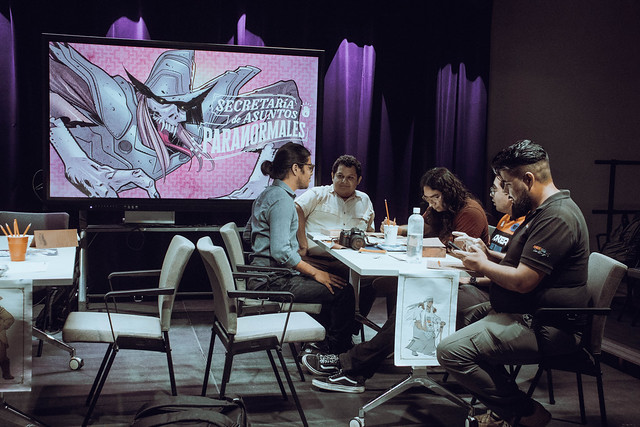 Cinco personas sentadas alrededor de una mesa jugando una partida de rol. Al fondo hay una pantalla con una ilustración del juego 'Secretaría de Asuntos Paranormales' desarrollado por la comunidad Leyendas del Noreste del Laboratorio Cultural Ciudadano de Monterrey (México).
