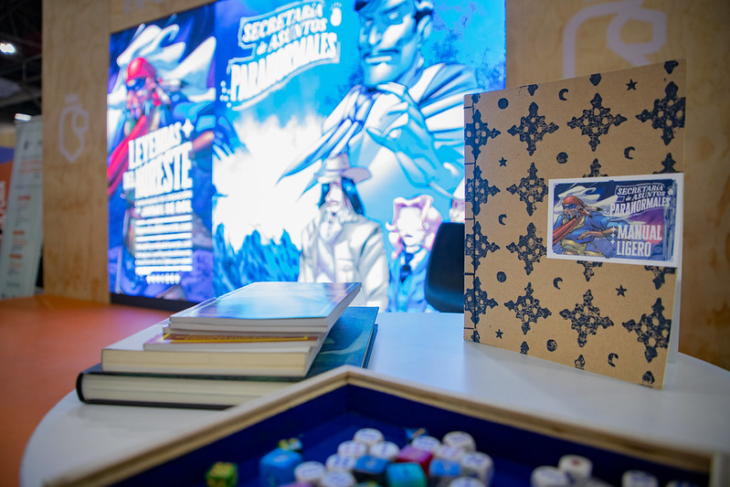 Escenario de la Feria Internacional del Libro de Monterrey con una pantalla con ilustraciones del juego de rol Secretaría de Asuntos Paranormales, en primer plano se ve un ejemplar del Manual Ligero sobre una mesa con otros libros y una caja de dados.