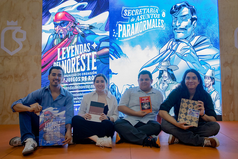 Parte del colectivo Leyendas del Noreste posa frente a una pantalla que proyecta ilustraciones del proyecto mientras sostienen material del juego de rol Secretaría de Asuntos Paranormales.
